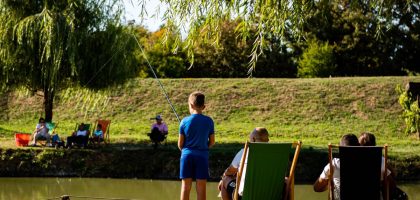 Young boy fishing by a pond on a sunny day, surrounded by trees and relaxed people.