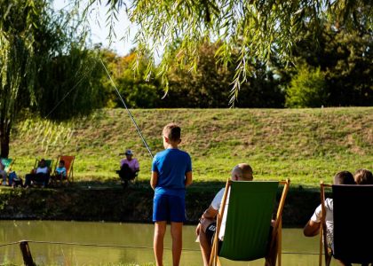 Young boy fishing by a pond on a sunny day, surrounded by trees and relaxed people.